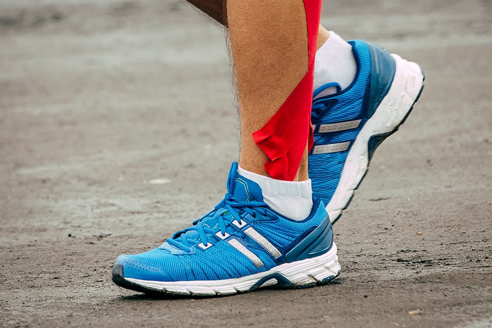 Onderbeen van hardloper met rode kinesiotape en hardloopschoenen.